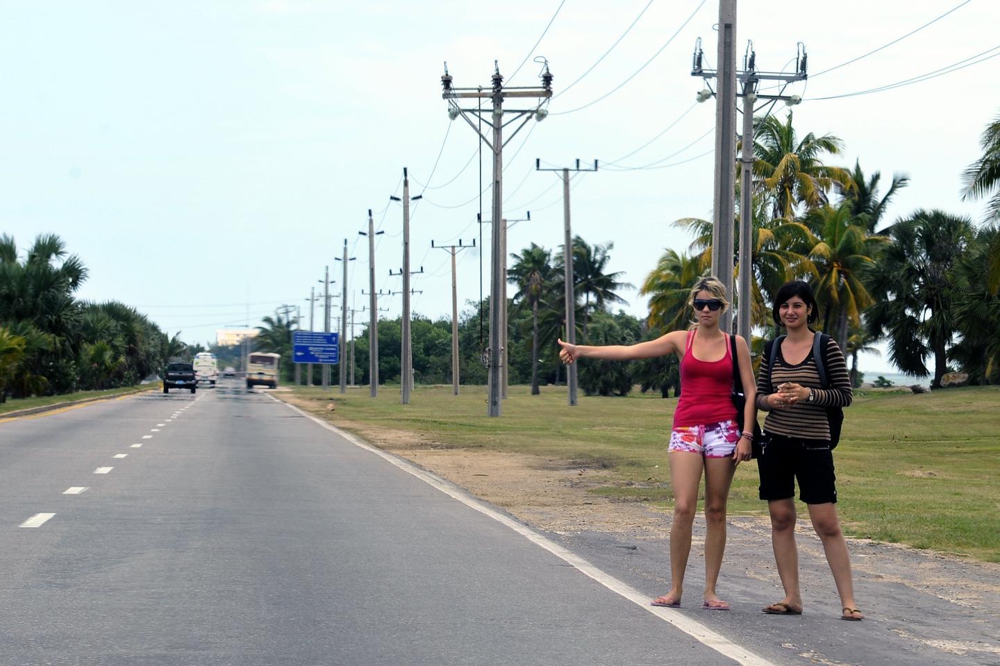 Jour. Extérieur. Vue de la route avec un panneau de signalisation, des camions en arrière-plan et deux femmes sur le côté, les mains tendues, arrêtant des voitures.